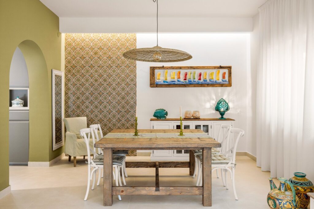 zona pranzo con parete decorata da piastrelle dell'epoca, tavolo in legno massello e sedie bianche in stile country chic, illuminata da una sospensione artigianale in corda natural