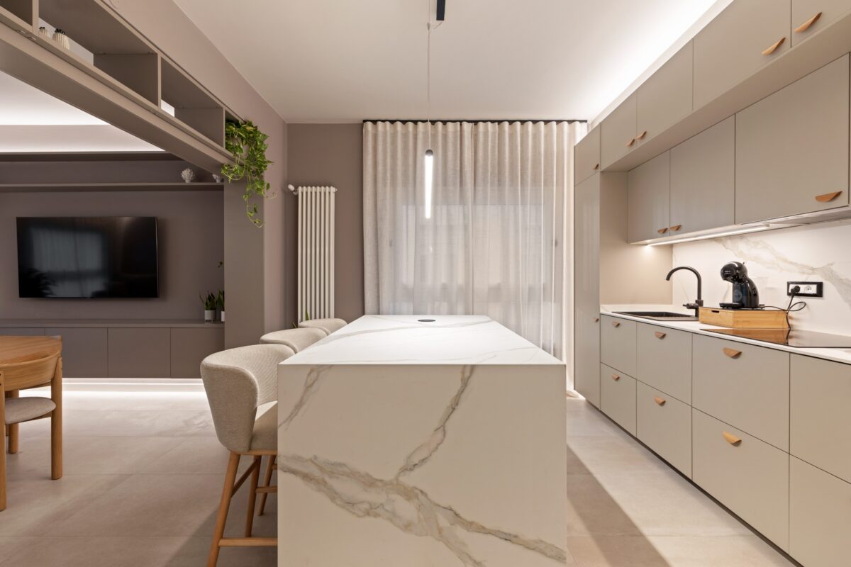Vista d'insieme della cucina con ante color tortora, isola centrale in marmo chiaro e zona living attrezzata in una struttura ricettiva di lusso a Palermo.